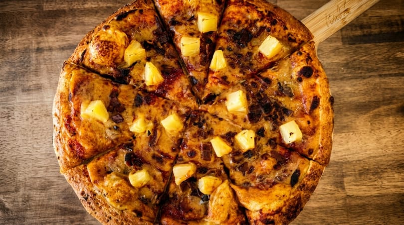 A sliced pineapple and bacon pizza on a wooden peel, viewed from directly above.