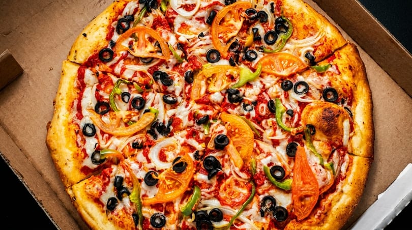 A freshly baked pizza with tomatoes, onions, olives, and green peppers in a cardboard box.